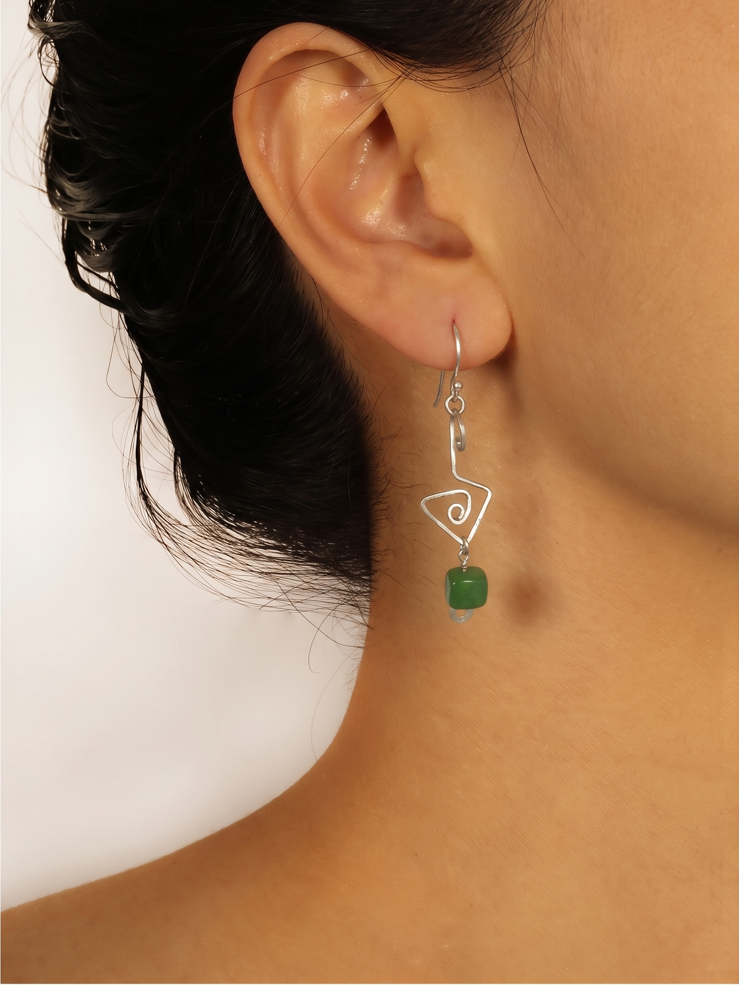 A close-up of a model's ear and neck wearing a silver dangle earring with a triangular outline and a small green aventurine accent.  Fish hook ear wires. Cheeky Goddess.