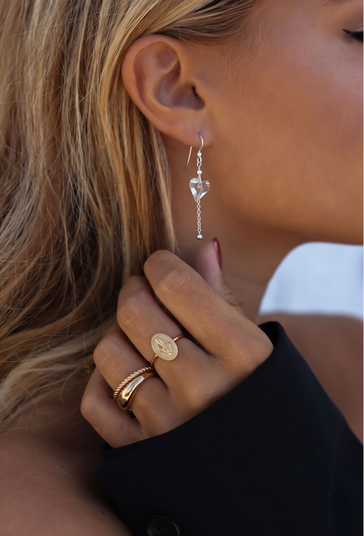 A close-up lifestyle shot of a woman wearing a delicate silver dangle earring with a clear heart-shaped crystal and a gold chain, along with gold rings on her finger.