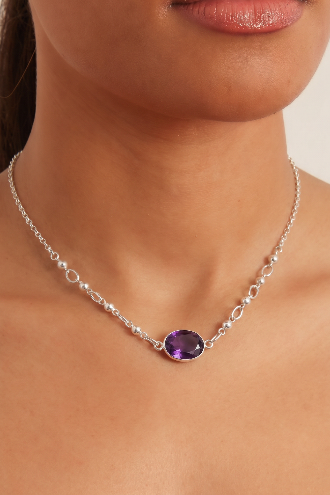 A close-up of a model's neck and chest wearing a delicate silver chain necklace with a polished oval purple gemstone pendant.