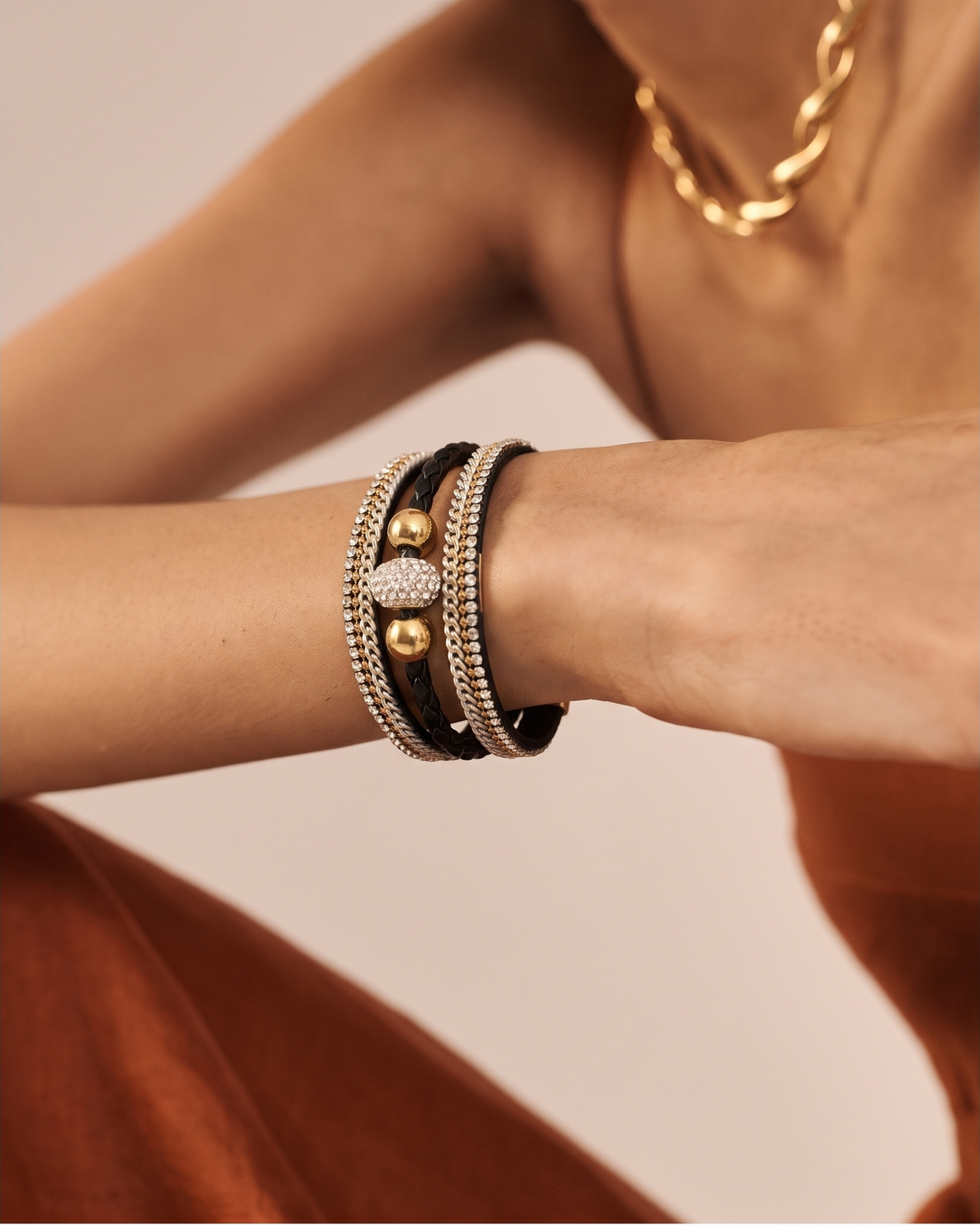 A close-up of a model's wrist wearing a stack of three bracelets: two black leather bands, one silver-toned beaded band, and a central gold-toned heart-shaped charm with a pavé diamond center. The model is wearing a rust-colored dress and a gold chain necklace against a soft beige background.
