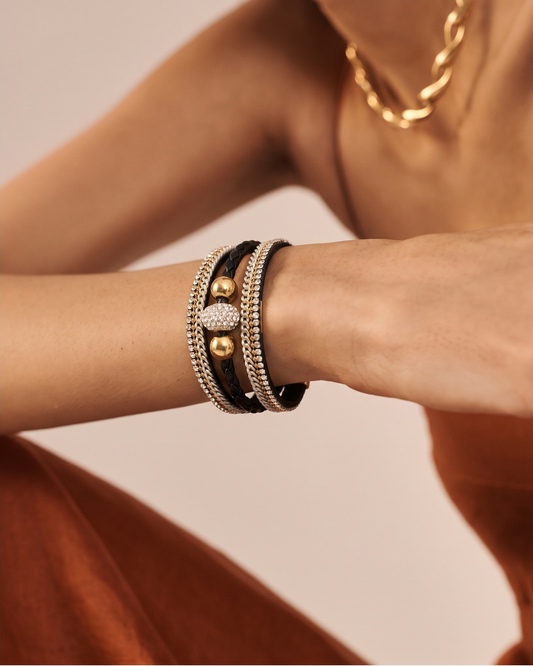 A close-up of a model's wrist wearing a stack of three bracelets: two black leather bands, one silver-toned beaded band, and a central gold-toned heart-shaped charm with a pavé diamond center. The model is wearing a rust-colored dress and a gold chain necklace against a soft beige background.