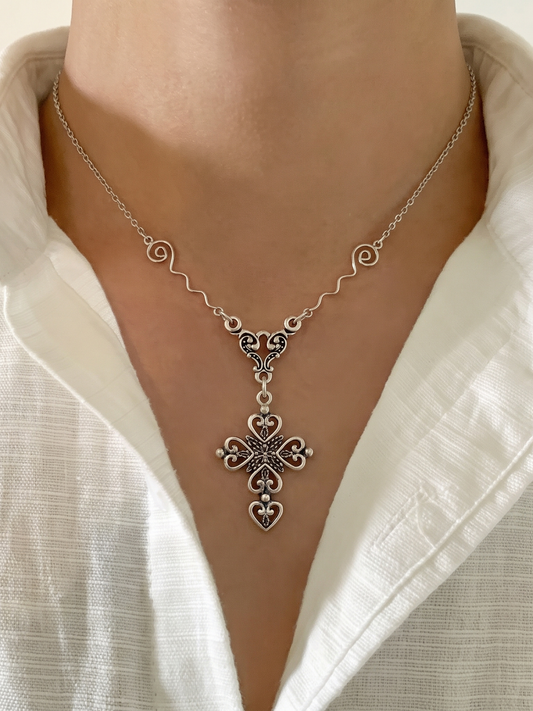 A close-up of a person's neck and chest wearing a silver-toned ornate cross pendant necklace over a white collared shirt.  Sterling silver. Cheeky Goddess.