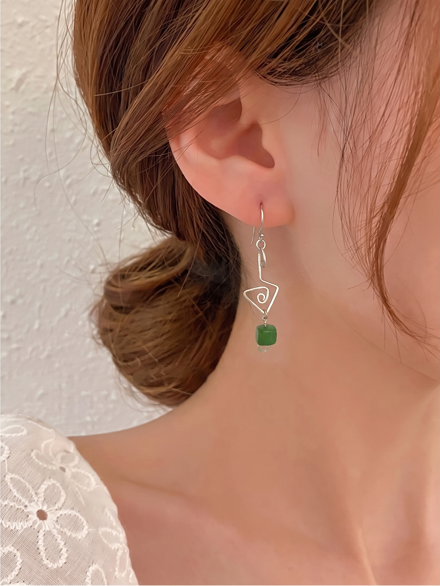 A close-up of a model's ear and neck wearing a silver dangle earring with a triangular outline and a small green aventurine accent.  Fish hook ear wires. Cheeky Goddess.